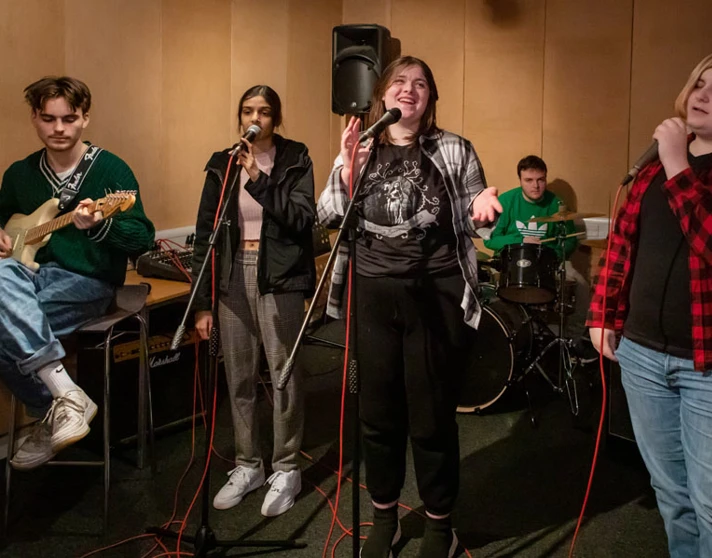 A group of five students rehearsing, featuring a guitarist, two vocalists, a bassist, and a drummer in a music studio. A group of five students rehearsing, featuring a guitarist, two vocalists, a bassist, and a drummer in a music studio.